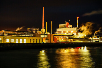 The Gersthofen industrial park at night. Industrial site with several buildings and tall chimneys, from which steam rises into the night sky. Reflection of artificial lighting in the Lech Canal.