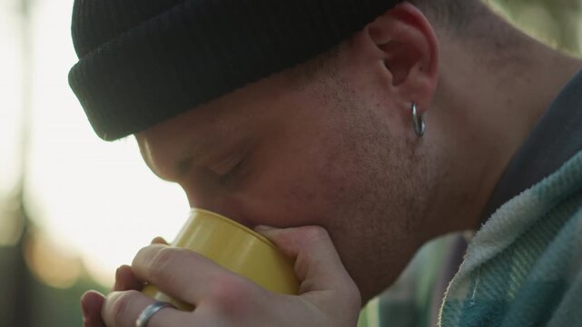 white hiker holding yellow mug closeup, sipping warm drink at forest campsite, hands with ring and worn fingers, wearing black beanie, soft morning sunlight through pines, introspective upward gaze