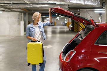 Naklejka premium A woman prepares to load her yellow suitcase into the trunk of her red car in a parking garage