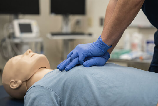Gloved hands are seen performing chest compressions on a mannequin with blue medical gloves visible in a clinical setting, highlighting the training process for cpr techniques