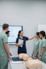 A group of medical trainees in green scrubs attentively listens to an instructor in navy scrubs who is explaining a procedure using cpr dummies in a bright training room with screens