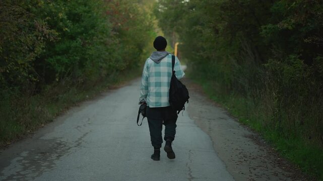 Photographer walking along country road carrying tripod and camera bag, plaid coat and sturdy boots on treelined lane, golden hour light and focused stride, location scouting for outdoor shoot
