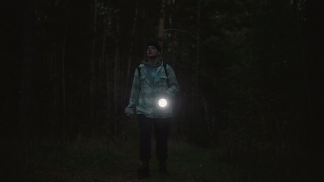 Caucasian wanderer night forest with flashlight casting beam on mossy path, tense atmosphere of darkness and shadow, looking skyward between trunks, isolated survival mood with faint glow and steady