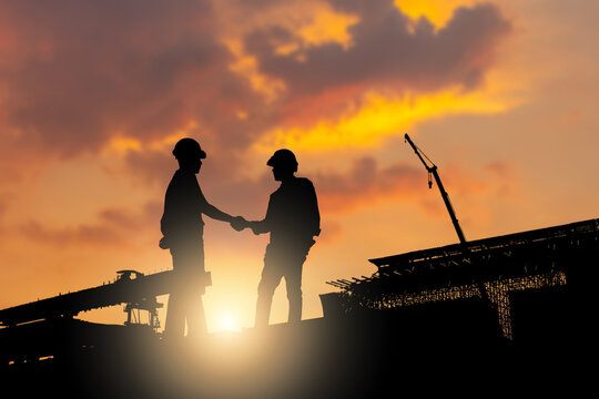 Silhouette of construction workers shaking hands at industrial site during sunset, Business partnership agreement between engineers at infrastructure construction site, Teamwork and success concepts