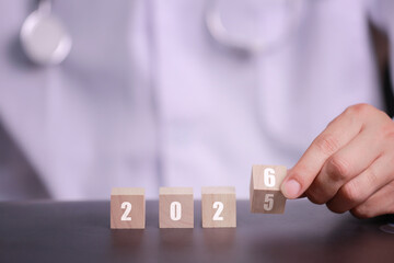 A hand holds a wooden block while rearranging numbered blocks displaying the years 2025 and 2026, suggesting a theme of time or planning.