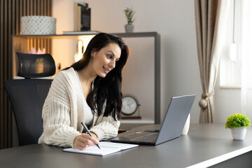 A woman attentively writes notes while participating in a live online session in her workspace.