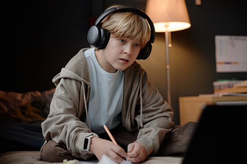 Caucasian child boy wearing headphones sitting on bed writing in notebook while looking at laptop screen showing focused expression representing young prodigy studying