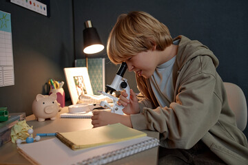 Caucasian boy child sitting at desk using microscope for scientific research, concentrating on examining specimen, surrounded by notebooks and study materials, demonstrating prodigy behavior