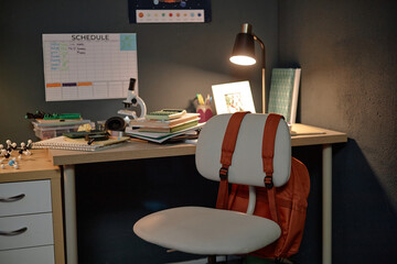 Desk with microscope, books, and science models, backpack hanging on chair, schedule and educational materials visible in background, no people present