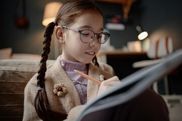 Caucasian girl child with long brown hair in braids wearing eyeglasses sitting on floor in bedroom writing in notebook, showing focused expression in cozy home study environment