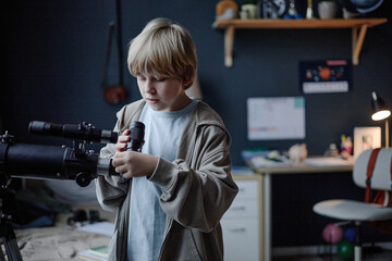Caucasian boy child adjusting telescope in room, demonstrating focus and curiosity, standing near desk with scientific materials, embodying young prodigy engaged in astronomy exploration © Seventyfour