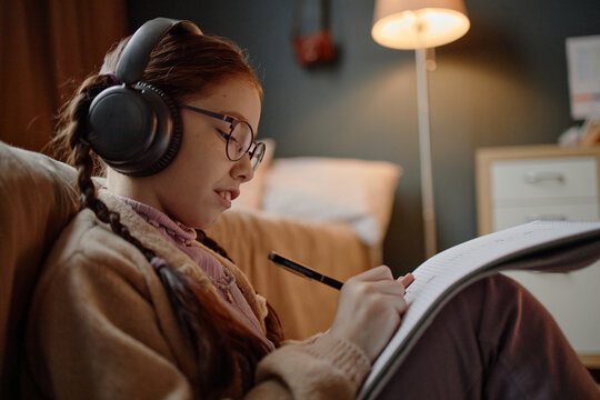 Caucasian girl child with long brown hair wearing glasses and headphones sitting on floor in bedroom writing in notebook, showing focused expression demonstrating prodigy talent