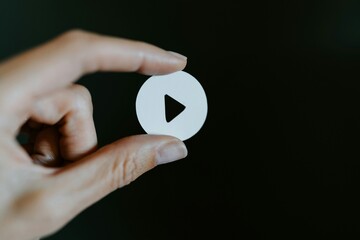A close-up, commercial shot of a person's fingers holding a round, white paper cutout of a play button icon against a dark black background, representing video content, media creation
