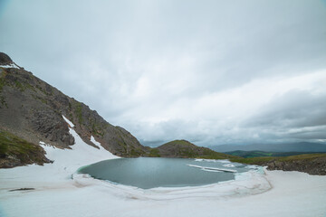 Alpine lake among snows in hanging valley with view to green hills far away under dramatic cloudy sky. Ice floes float in glacial lake in mountain cirque against hilly vastness in gloomy rainy weather