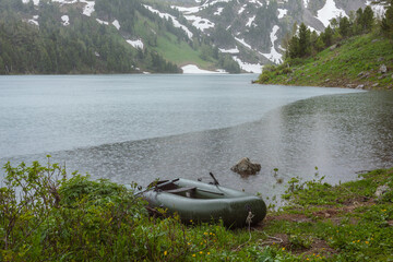 Inflatable boat with oars among lush thickets on woody shore of alpine lake with view to conifer forest hills and rocky snowy mountain wall during heavy rain. Circles on water surface when it rains.