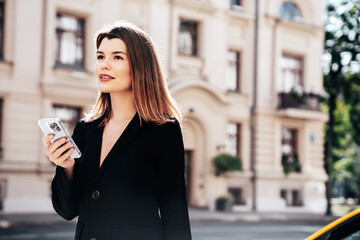 Fototapeta premium Young beautiful woman in nice trendy white long jacket suit. Sexy smiling model posing in street. Fashionable female outdoors. Cheerful and happy. Holds smartphone, uses phone apps, looks at screen