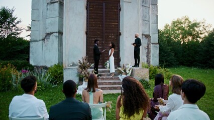 Fototapeta premium The groom reads the wedding vow standing in front of the bride near the ancient church. Wedding ceremony with guests and a priest