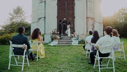 The bride and groom smile looking at each other standing in front of the priest at an outdoor wedding