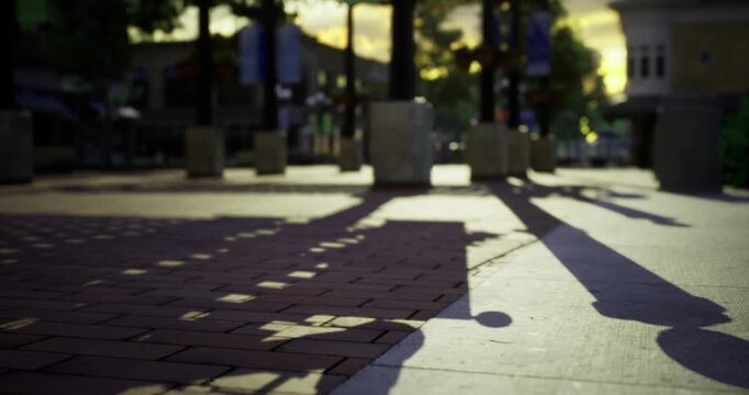 Warm sunset light filters through tall buildings, casting elongated shadows on the cobblestone sidewalk. A tranquil moment in a bustling urban environment, filled with evening possibilities.