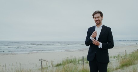 The groom smiles looking at the camera and adjusting cufflinks on his sleeve while standing on the...