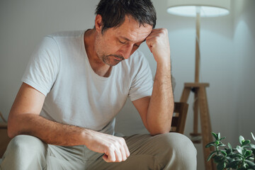 Sad mature man sitting in living room near floor lamp, leaning on fist and staring down with emotional exhaustion, loneliness and depression mood in dim cozy interior.