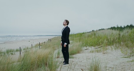 A man in formal clothes straightens his hair standing on the seashore and looking into the distance...