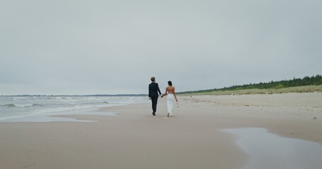 The bride and groom hold hands walking on the beach, the bride holds shoes in her hands