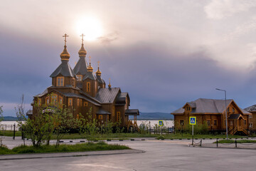 Holy Trinity Cathedral in Anadyr, Chukotka, Russia, wooden Orthodox church with golden domes overlooks road and bay under cloudy sky.
