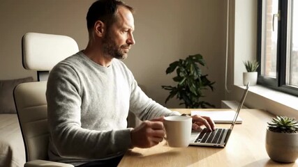 Focused adult man working intently on a laptop computer while enjoying a warm beverage at his well-lit home office desk indoors