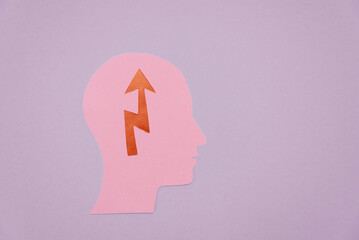 A pink paper cutout of a human head silhouette with an upward arrow and zigzag pattern inside. The background is a soft lavender color.