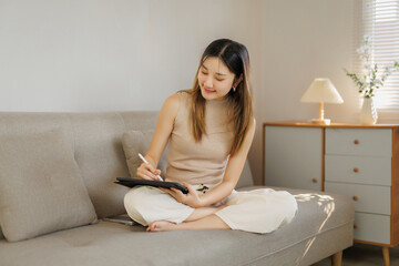 Young Asian woman sitting on a beige sofa, engaging with a digital tablet using a stylus for creativity and art
