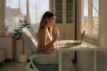 Young woman smiling and waving during a video call on a laptop, working remotely from a sunlit home office
