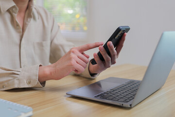 Person using mobile phone for communication, browsing, or online activity with a laptop open on a wooden desk