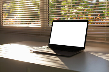 Laptop with blank white screen on a bright desk by a window with blinds, showing warm sunlight and a home office setup