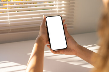 Woman hands holding a mobile smartphone with a blank white screen, standing near a window with blinds and sunlight