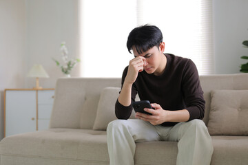 Young man sitting on a sofa, looking upset while holding a smartphone, experiencing headache and feeling disappointed
