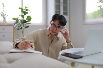 Young man with glasses focused on brainstorming and writing in a notebook while working remotely at a laptop in a home office