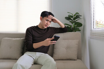 Young Asian man looking disappointed while holding a smartphone, experiencing stress or bad news on a sofa at home