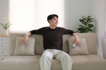 Smiling young man sitting on a comfortable sofa in a modern living room, looking away while relaxing at home