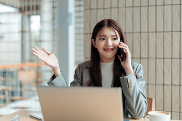 Smiling young Asian business woman talking on mobile phone, holding a pen, working on laptop in a modern office space