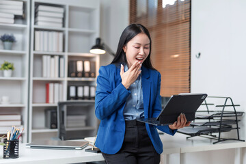 Young businesswoman yawning, looking tired and covering her mouth with hand while working on a digital tablet in office