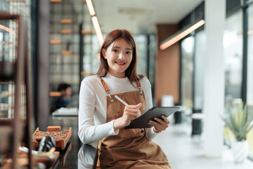 Young Asian woman wearing apron, smiling and typing on a tablet, managing her small business in a modern cafe