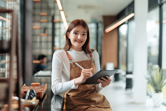Young Asian woman wearing apron, smiling and typing on a tablet, managing her small business in a modern cafe - Powered by Adobe