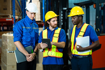 Warehouse workers wearing helmets and reflective vests standing inside warehouse discussing logistics operation using tablet and holding clipboard promoting teamwork coordination professional