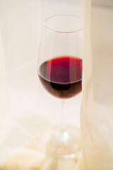 Fototapeta premium A wine glass filled with dark red liquid is partially visible behind soft, flowing curtains