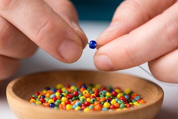 Woman hands stringing a blue bead onto a needle above a bowl of colorful beads. Creating jewelry for hobby and craft.