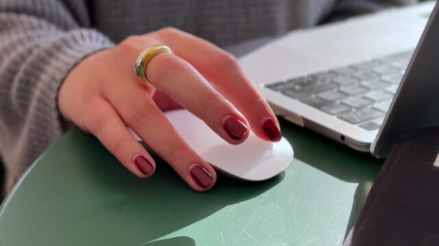 Close-up shot of a hand with red polished nails and a gold ring using a wireless mouse next to a laptop on a desk&mdash;ideal for tech, business, or remote work-related visuals.