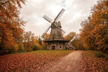 Old windmill building surrounded by autumn trees and fallen leaves in a rustic countryside landscape. Traditional architecture on a fall day.