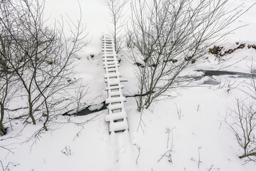 Small Wooden Bridge in Winter Snowy Landscape Over a Stream - Untouched Nature After Snowfall, Aerial View.