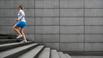 Beautiful middle-aged woman running up concrete stairs against background of concrete wall in city on summer day. Side view 